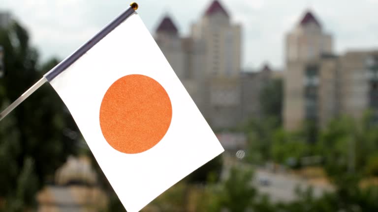 Flag of Japan set against blue sky and city street. Japan flag waving. The holiday flag of Japan illuminated by the sun. White banner bearing a crimson-red circle at its center. Copy space for text