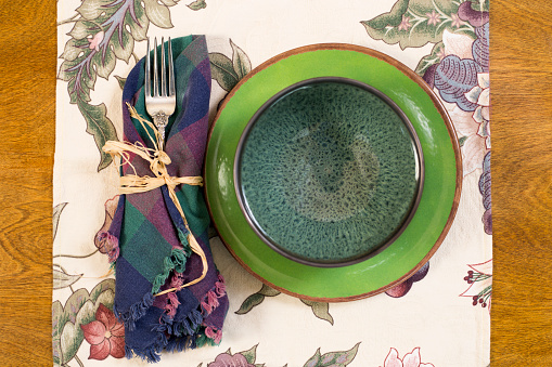 Food and Drink - Holiday Season - Place Settings.  Plaid napkin tied with raffia, green plate with blue bowl on table runner, oak table.