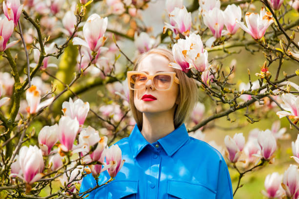 stilvolle frau in brille und blauem hemd in der nähe von magnolienbaum im frühling - magnolia blossom flower head single flower stock-fotos und bilder