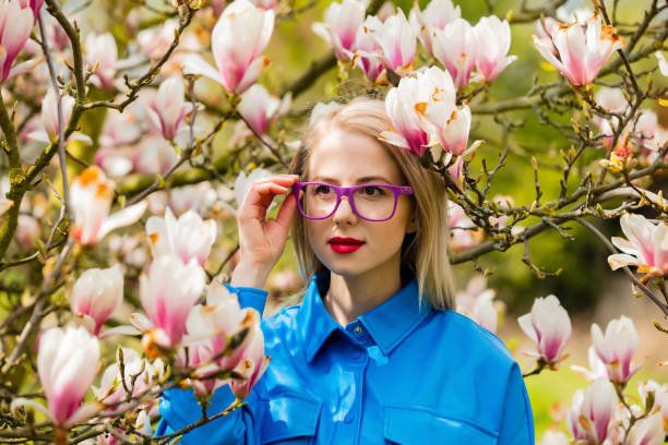 stilvolle frau in brille und blauem hemd in der nähe von magnolienbaum im frühling - magnolia blossom flower head single flower stock-fotos und bilder