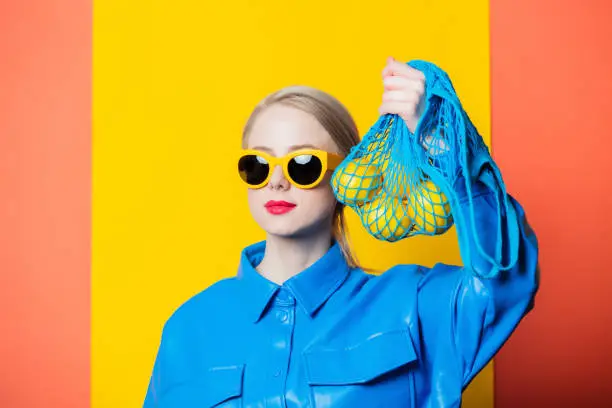 Photo of Stylish woman in blue shirt and yellow sunglasses with lemons in net bag on yellow and coral color background