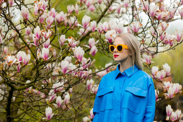 schöne blonde frau mit sonnenbrille und blauem hemd neben magnolienbaum im frühling - magnolia blossom flower head single flower stock-fotos und bilder