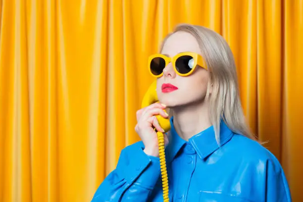 Photo of Stylish woman in blue shirt and yellow sunglasses stands with retro dial phone on yellow curtains background