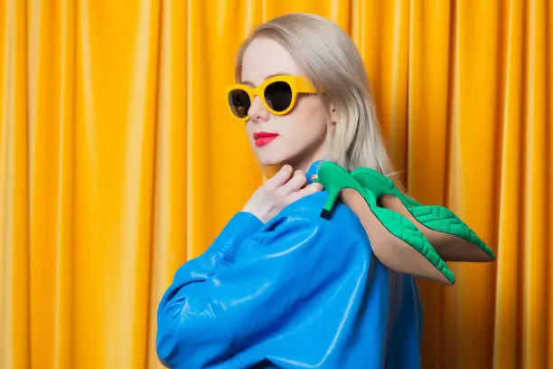 Photo of Stylish woman in blue shirt and yellow sunglasses hold heels on yellow curtains background