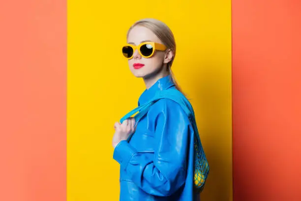 Photo of Stylish woman in blue shirt and yellow sunglasses with lemons in net bag on yellow and coral color background