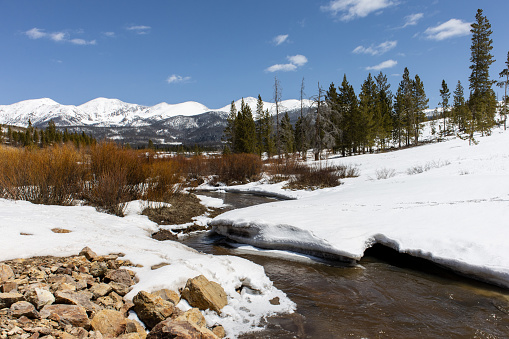 A river runs through the Rocky Mountains of Colorado in the winter with snowy river banks and mountains in the background