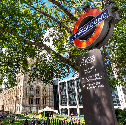 London,England,UK-August 21 2019: The Underground sign,at top of steps,leading down to a five minute underpass walk to South Kensington tube station, visitors sit on the grass,in fine sunshine.