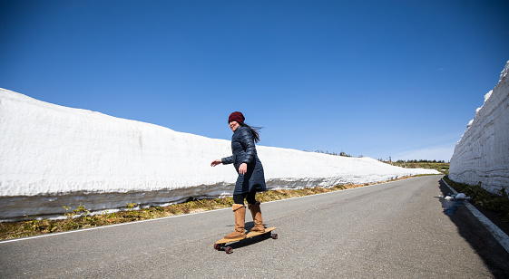 One woman skateboarding down a mountain road with no cars. The sides of the road have tall walls of snow.