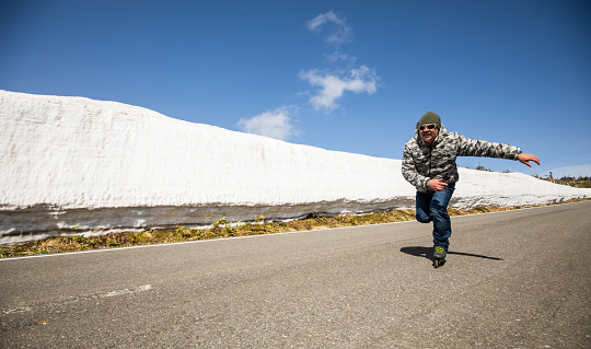 Man inline skating past low angle camera along a snowy mountain road with snow walls on the side of the road.