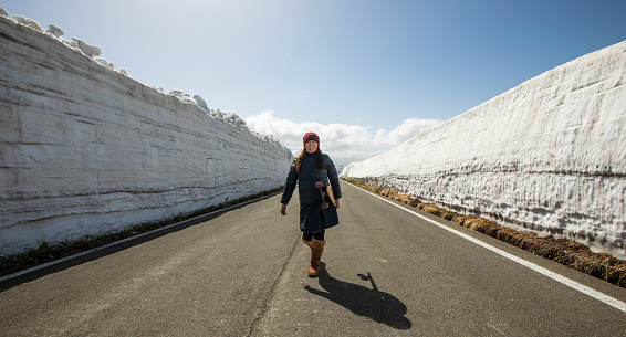 One woman with her skateboard walking down a mountain road with no cars. The sides of the road have tall walls of snow.