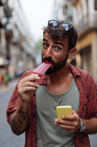 Smiling young man eating ice cream and using his phone while walking through a city street