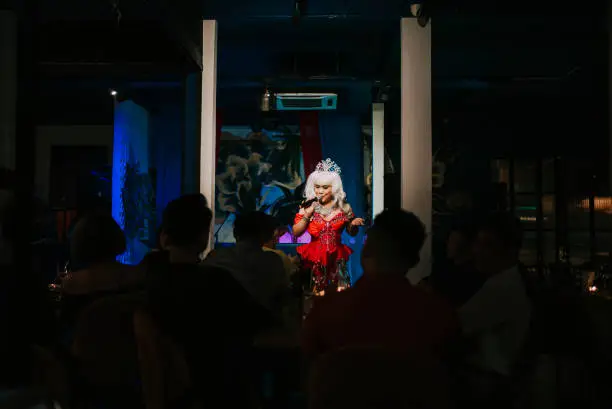 Photo of Asian drag queen in spot lit stage performance at pub with audience in silhouette