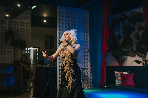 Asian drag queen in spot lit stage performance at pub with audience in silhouette