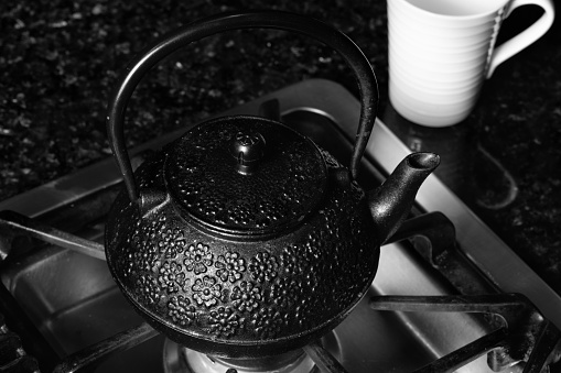 Cast iron tea pot on a stove and a white cup. Black and white photograph.