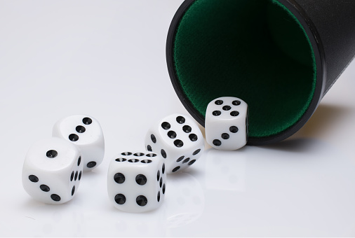 Close up of black cup tumbler mug with green felt cover and five white dice rolling out of it showing different numbers on white background as concept for dice poker and gambling