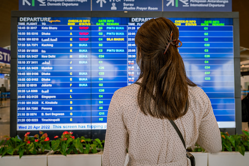 Back side of young Asian businesswoman standing over the flight board for check-in at the flight information screen in modern an airport, travel and transportation with technology concept.