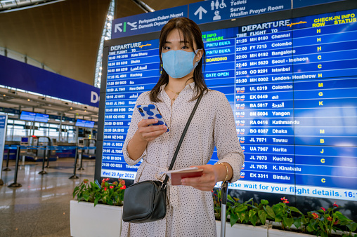 An Asian female business wearing a facemask holding passport using smartphone checking her boarding schedule at the airport. Looking at camera. Individuals Business Travelling Concept.