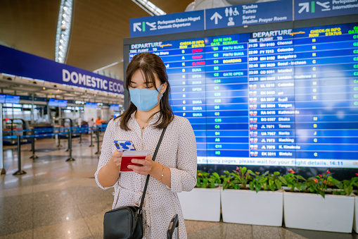Female traveler wearing a facemask at the airport holding passport while texting on her phone. Individuals Business Travelling Concept.