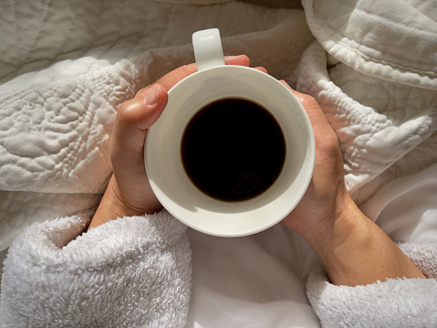 Top view of an asian woman holding a white coffee cup of espresso in the early morning sunshine surrounded by blankets