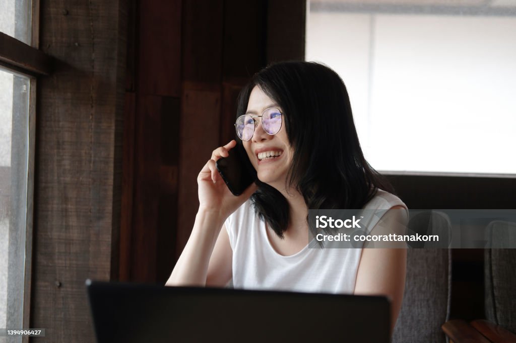 Smiling young Asian woman is wearing glass and calling with smartphone and online working with laptop in living room. Work from home for Covod-19 outbreak concept. Adult Stock Photo