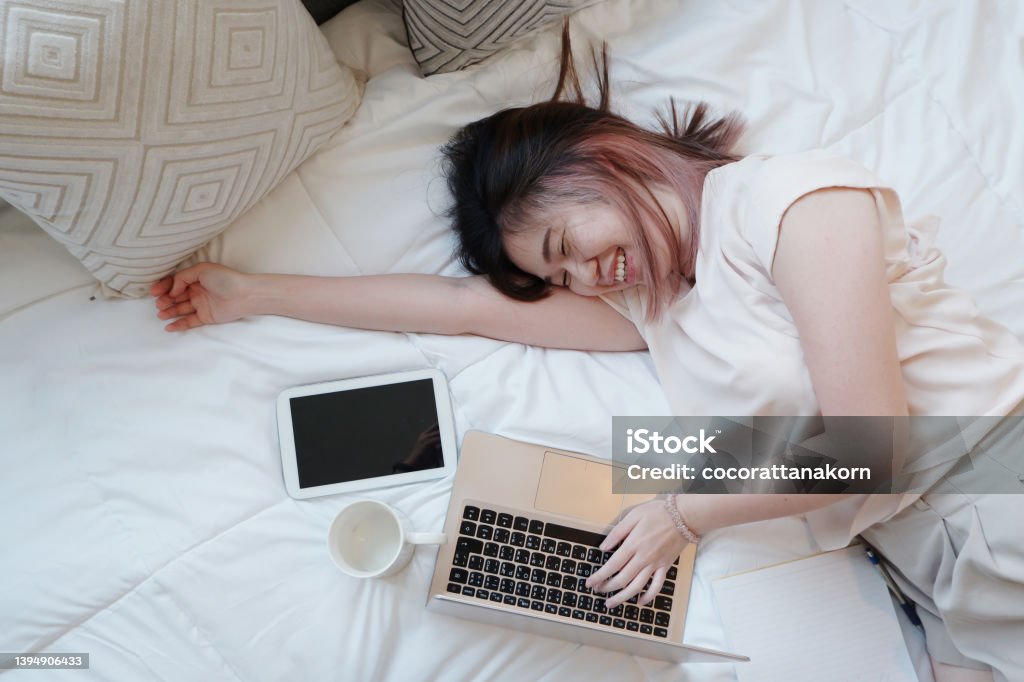 Smiling Asian businesswoman is relaxing and laying with laptop in bedroom on holiday. She is online working from home with technology during the Covod-19 outbreak concept. Adult Stock Photo