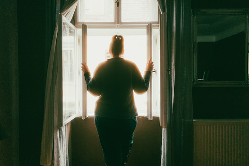 Color image depicting the rear view of a lonely woman looking at the street from her bedroom window. She is rendered an almost total silhouette due to the light flooding into the room from the window. Room for copy space.