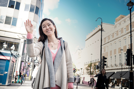 Smiling oriental mature woman waving someone in the street. London busy street.