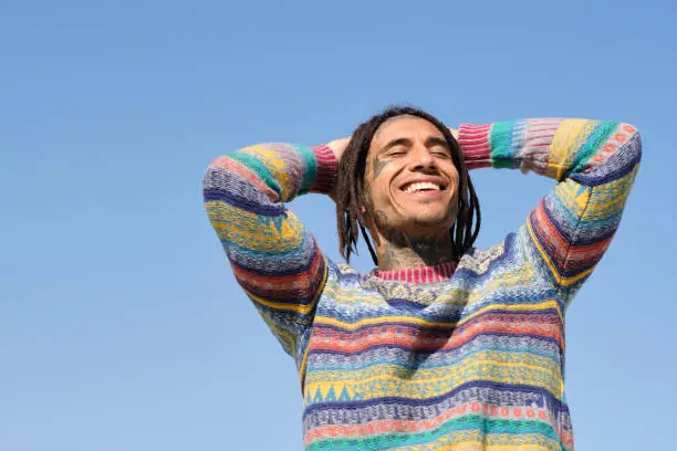 happy handsome young face tattooed Caribbean man with a blue sky and arms on his head and eyes closed