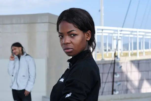 profile portrait of a millennial black woman looking intensely at the camera and a guy in the background talking on the phone
