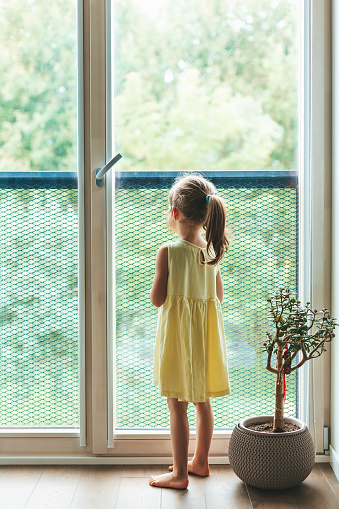 Rear view of a little girl in yellow dress standing near the window and looking through it