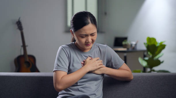 Asian woman was sick with chest pain. Asian woman using hands touching her chest was sick with chest pain sitting on sofa in living room at home. long covid stock pictures, royalty-free photos & images