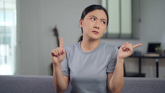 Asian woman feel confused shrugging and pointing up sitting in living room at home.