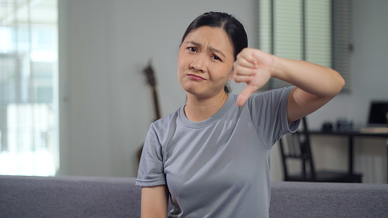 Asian woman annoyed looking at camera showing thumb down and sitting on sofa in living room at home.