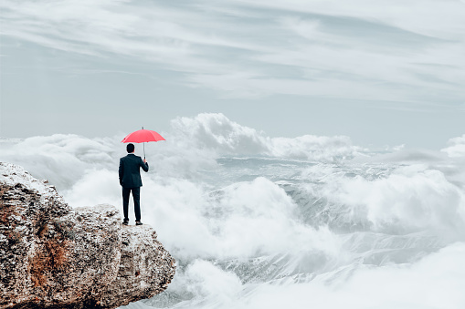 A businessman stands on top of a mountain top as he holds an umbrella as he tries to protect himself from risk.