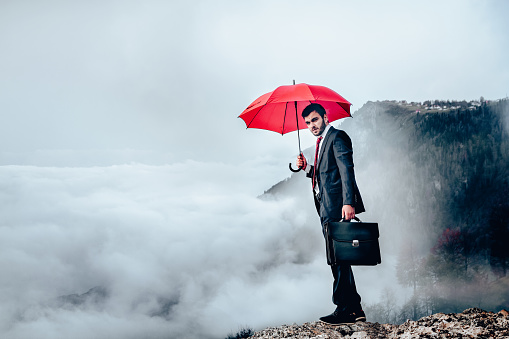 A businessman stands on top of a mountain top as he holds an umbrella as he tries to protect himself from risk.