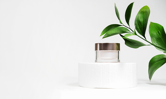 A mock up white jar of cream on a white gray background, on a white table with green leaves of a tropical plant, with hard shadows. Stylish look of the product, identity. Cylindrical podium
