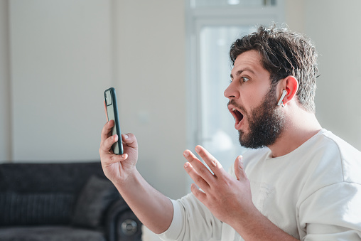 Portrait of shocked young man looking at smart phone's screen receiving bad news at the home. Horizontal composition.