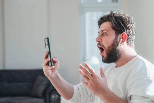 Portrait of shocked young man looking at smart phone's screen receiving bad news at the home stock photo