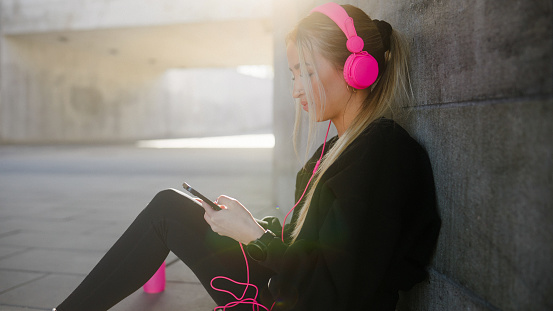 A female jogger, sitting by a wall in a concrete underpass on a sunny day. She is some music on her smartphone for motivation.
