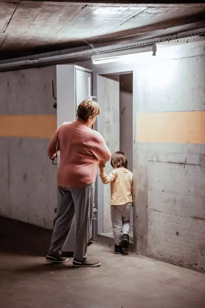 Senior woman with little child entering nuclear fallout shelter built in basement of building opening thick metal hermetic door
