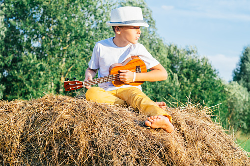 Portrait of barefoot boy in hat on haystack in field. Playing small guitar, ukulele. Light sunny day. Cheerful and music concept. Outdoor activity. Trees on background, Countryside