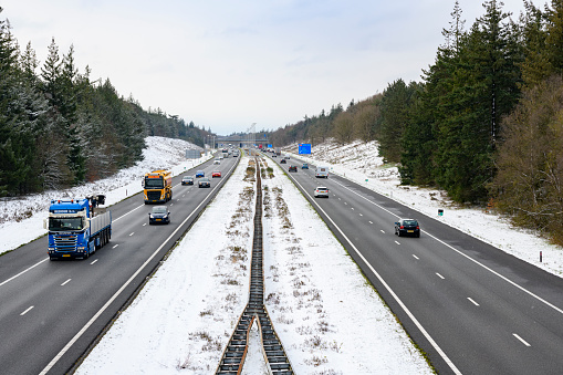 Highway through a snowy forest landscape with colorful cars and tracks driving on the black asphalt. The A50 highway through the Veluwe nature reserve is surrounded by snow covered pine trees.