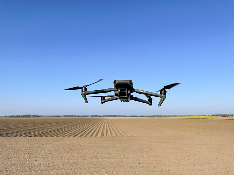 DJI Mavic 3 drone flying in the air over agricultural fields with the Hasselblad gimbal camera pointing forward. The Mavic 3 is a teleoperated quadcopter drone for aerial photography and videography mad by Chinese manufacturer DJI.