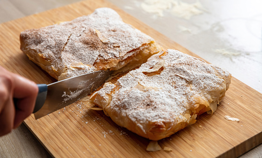 Greek pastry with cream sugar powder and ground cinnamon. Hand with a knife cutting bougatsa, traditional custard pie on a wooden board, close up view.