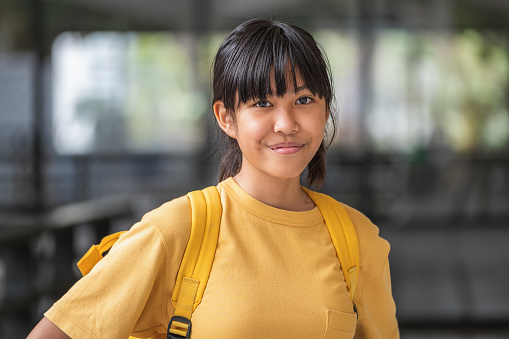 Portrait of young restrained smilling Asian girl wearing yellow t-shirt and backpack standing at school outdoors, Happy Thai teenage student girl wearing casual clothing and looking at camera