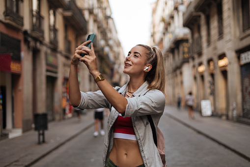 Photo of a happy young woman taking pictures with her smartphone while exploring a city