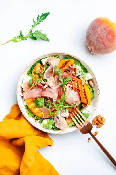 Summer salad bowl with sweet grilled peach, jamon, soft cheese, walnuts and fresh arugula on white kitchen table background, top view, negative space