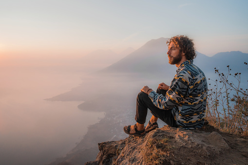 Young Caucasian man sitting on mountain on  the background of Atitlan Lake in Guatemala at sunset