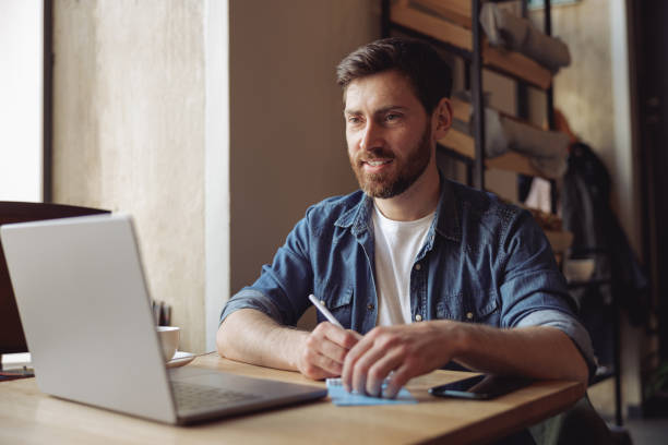 istock Handsome smiled man taking notes in notebook and working in cafe at laptop. Freelancing job. 1394954791