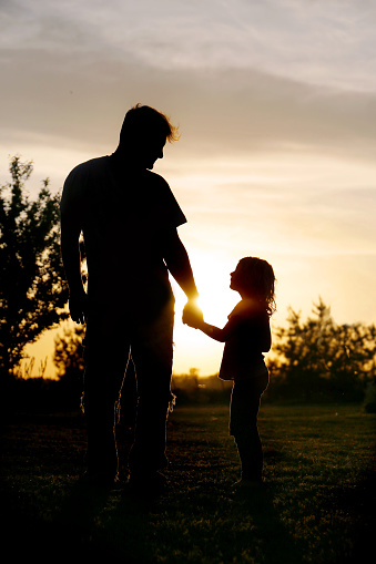 A silhouette of a loving father holding hands with his little daughter child outside while enjoying the sunset.